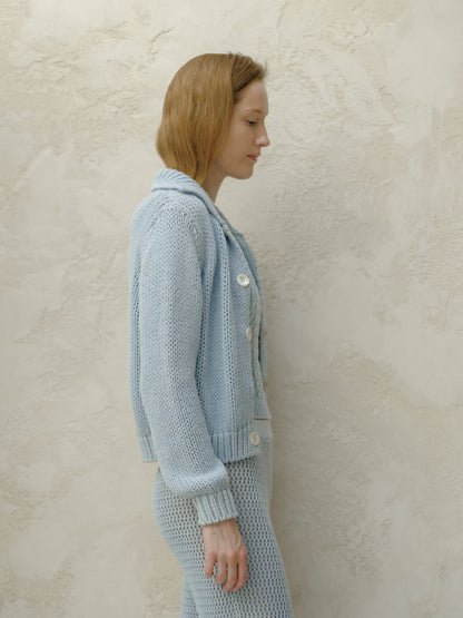 Side profile of a woman in a light blue knitted bomber jacket with white buttons against a textured beige wall.