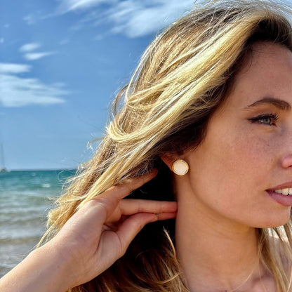 Camelia white ceramic stud earring with gold luster worn on a woman's ear at the beach