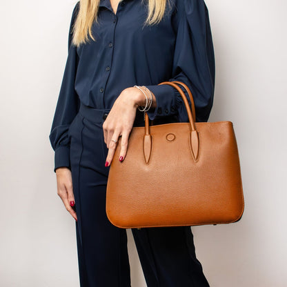 Camelia Italian leather tote bag in tan pebbled leather with two top handles held by a model in a navy outfit.