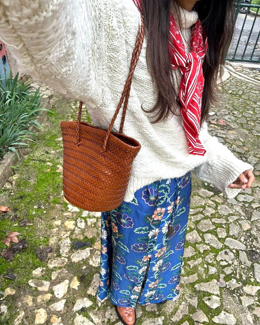 Carmel hand woven brown leather shoulder bucket bag worn on the shoulder with a braided strap, paired with a cream knit sweater and red scarf.