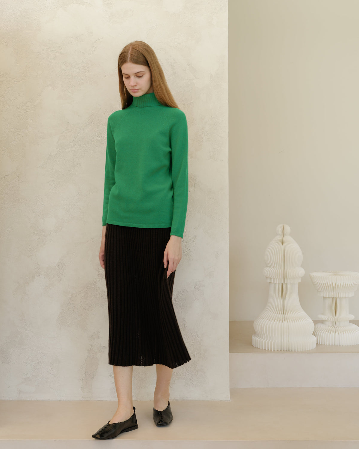 Modelo con verde intenso knit jersey de cuello alto and black midi falda plisada against a textured beige wall.