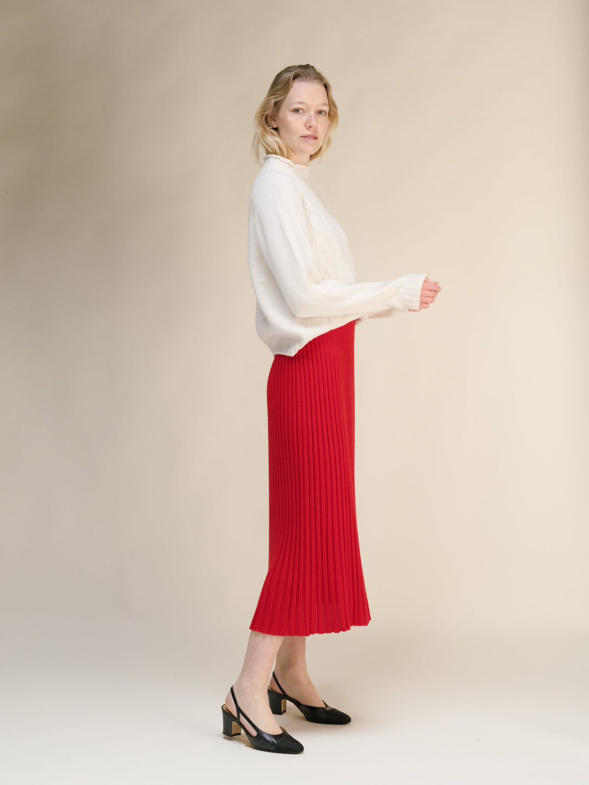 Side profile of a modelo con a cream knit sweater and red falda midi acanalada with black heels.