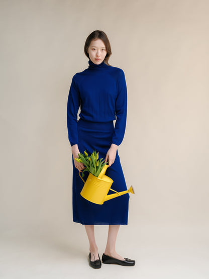 Model in a royal blue turtleneck dress holding a yellow watering can with green plants against a neutral backdrop