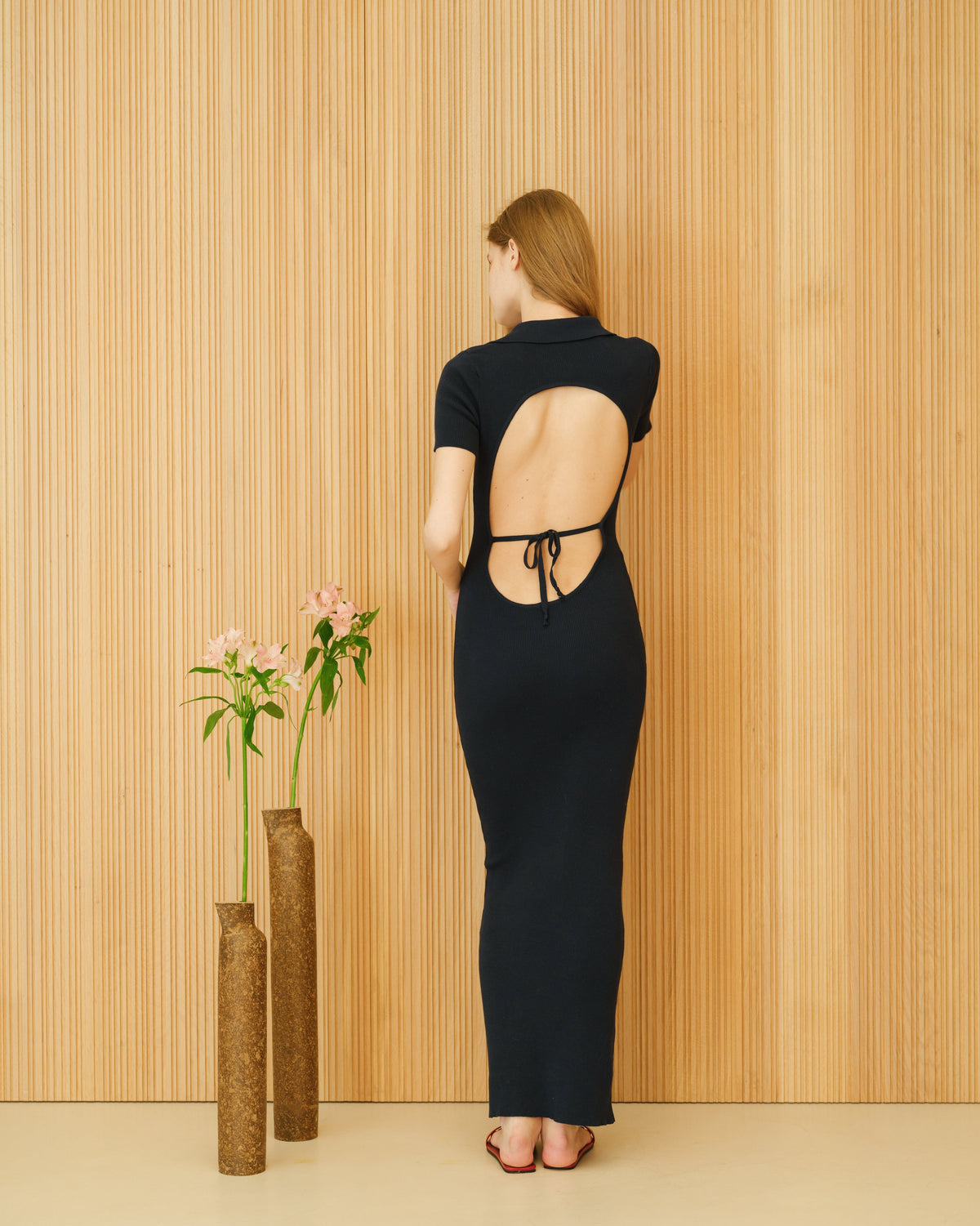 Back view of a model wearing a black open-back cotton dress with tie strings standing against a wood panel wall.