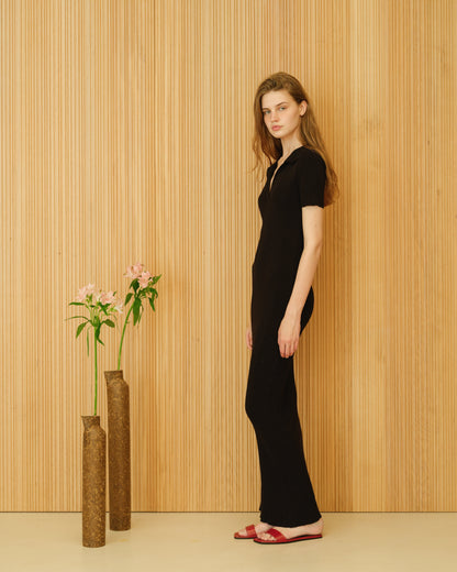 Model in a black cotton dress stands in profile against a wood panel wall with brown vases and pink flowers.