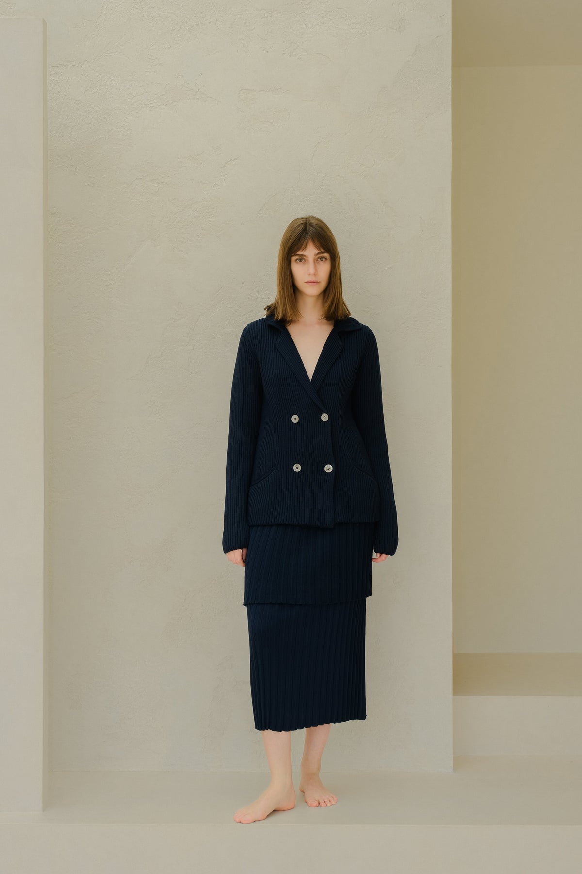 Azul marino knitted double breasted blazer with a layered falda larga plisada on a barefoot model against a beige textured wall