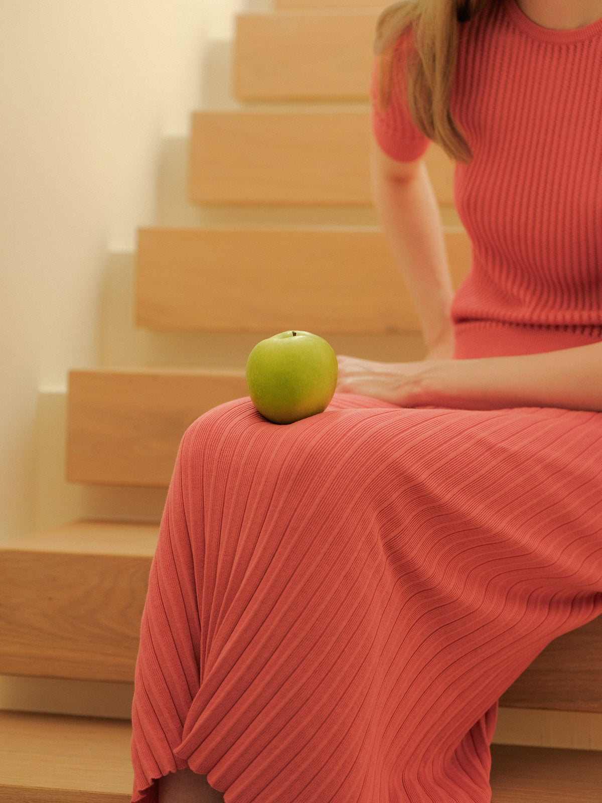 Verde apple rests on a coral pink long falda plisada llevado por una modelo sitting on wooden stairs.