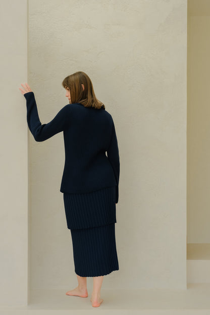 Back view of a modelo con a azul marino knit plissé skirt set against a beige wall.