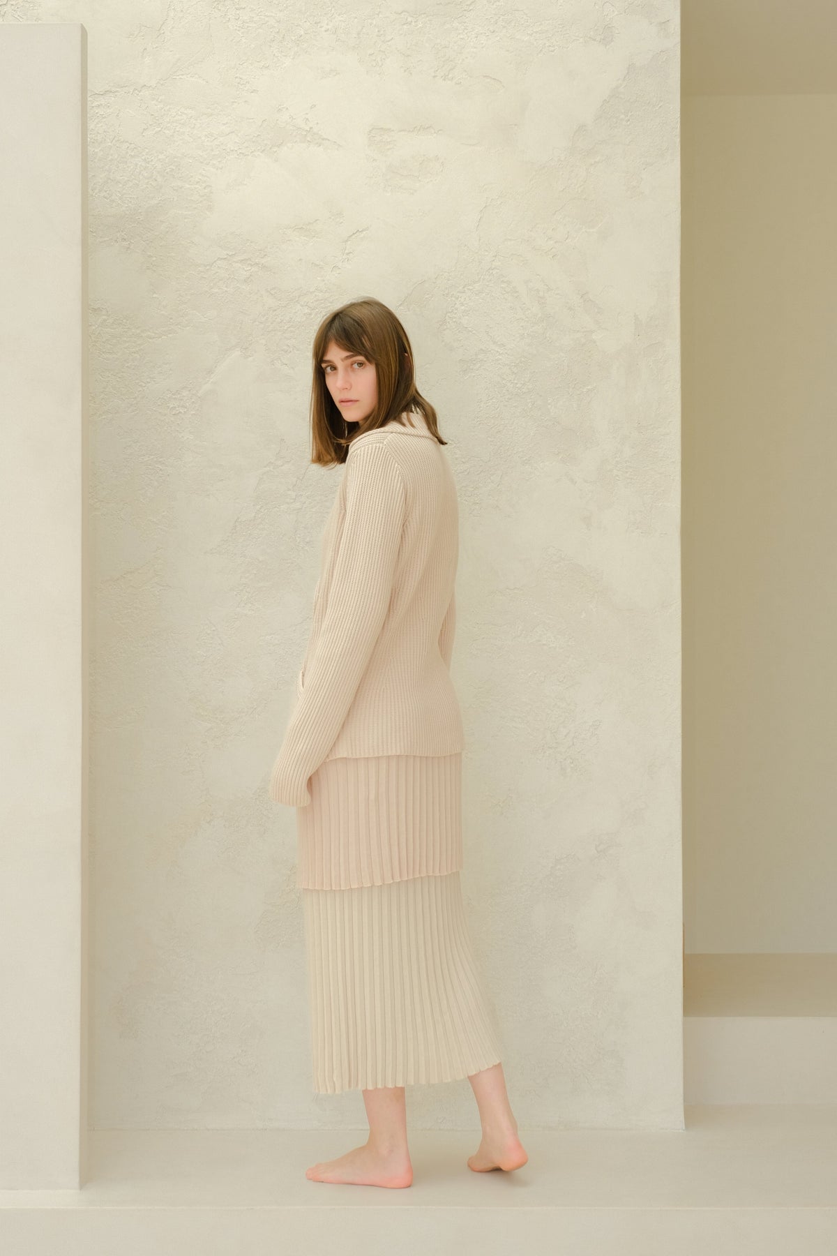 Vista lateral of a model in a beige Fine Cotton Plissé knit ensemble with a long falda plisada