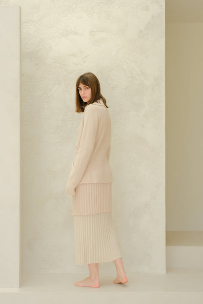 Vista lateral of a model in a beige Fine Cotton Plissé knit ensemble with a long falda plisada