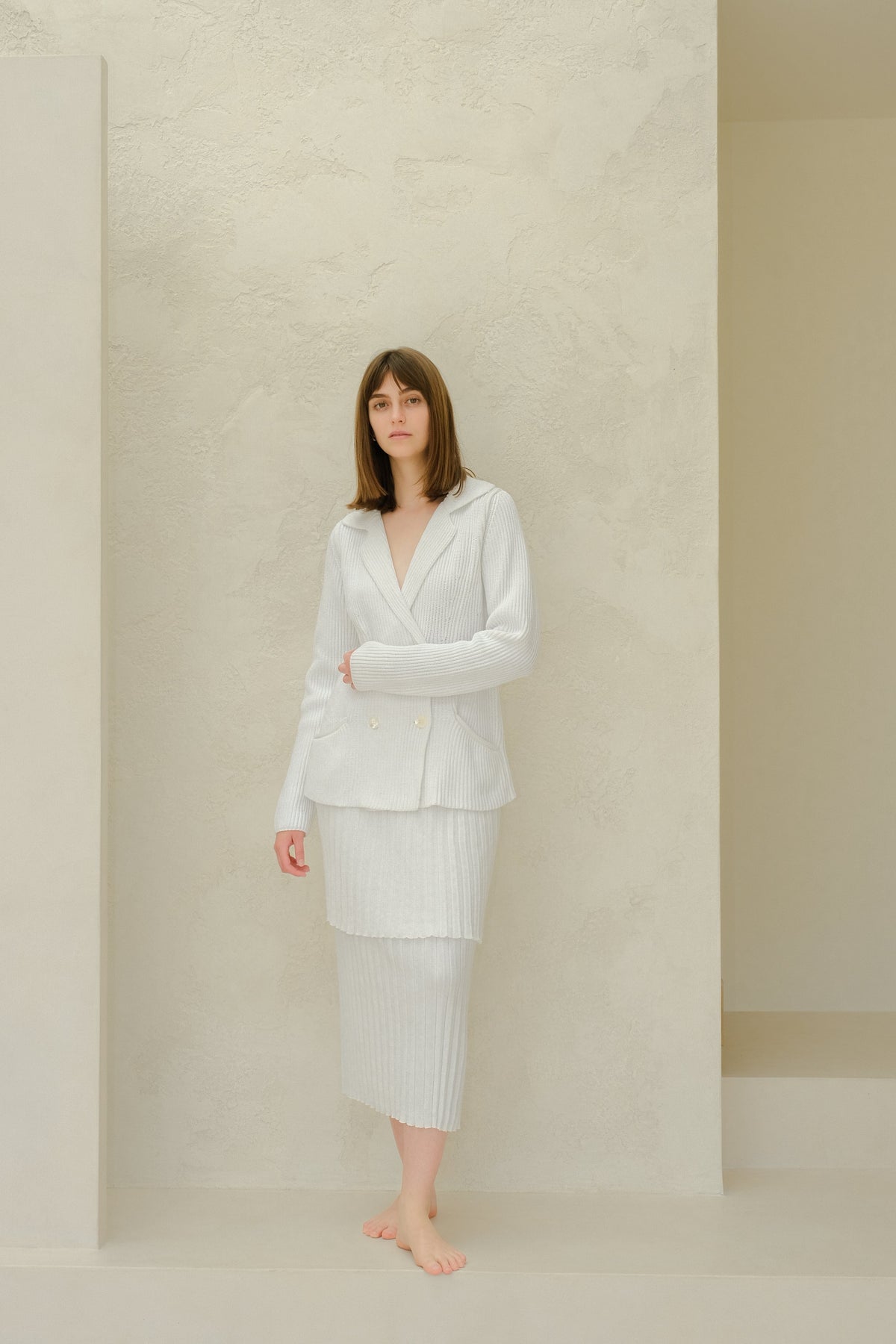 Vista lateral of the model in a pale gray blazer de punto and falda larga plisada a juego against a textured light wall.