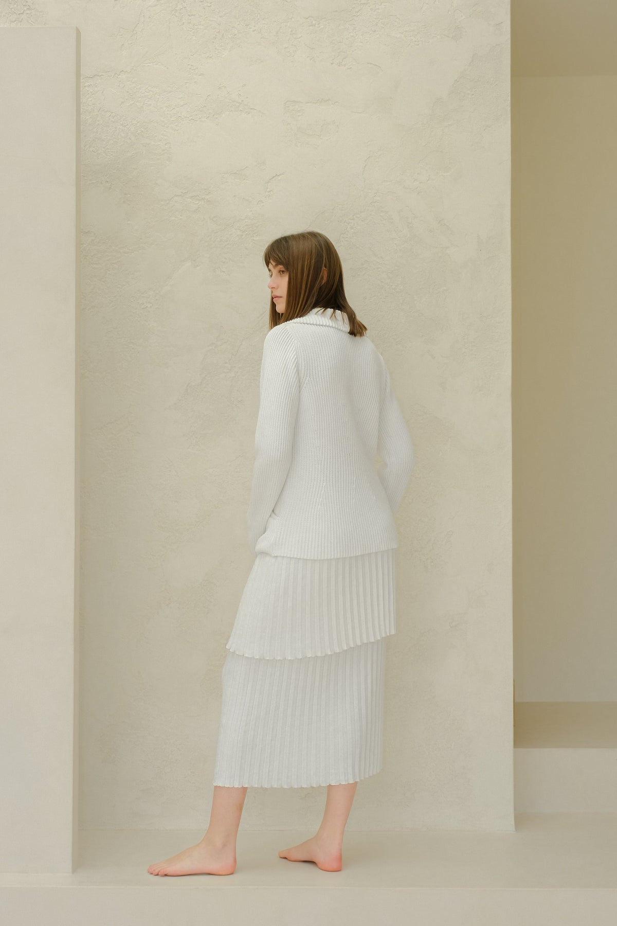 Vista lateral of a modelo con a white fine cotton plissé knit skirt with a jersey a juego against a textured beige wall.