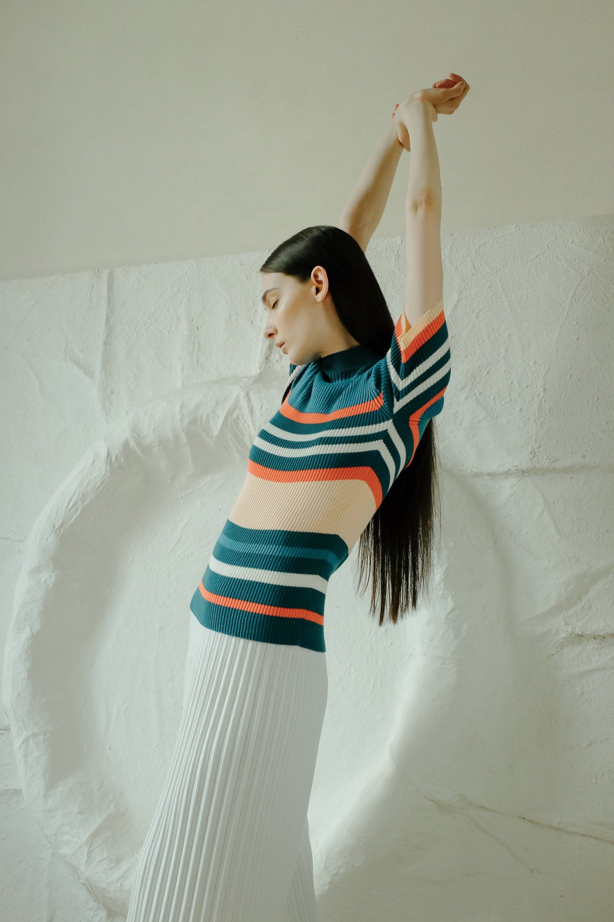 Profile of a model in a multicolor striped knit top and a white falda larga plisada, arms raised overhead.
