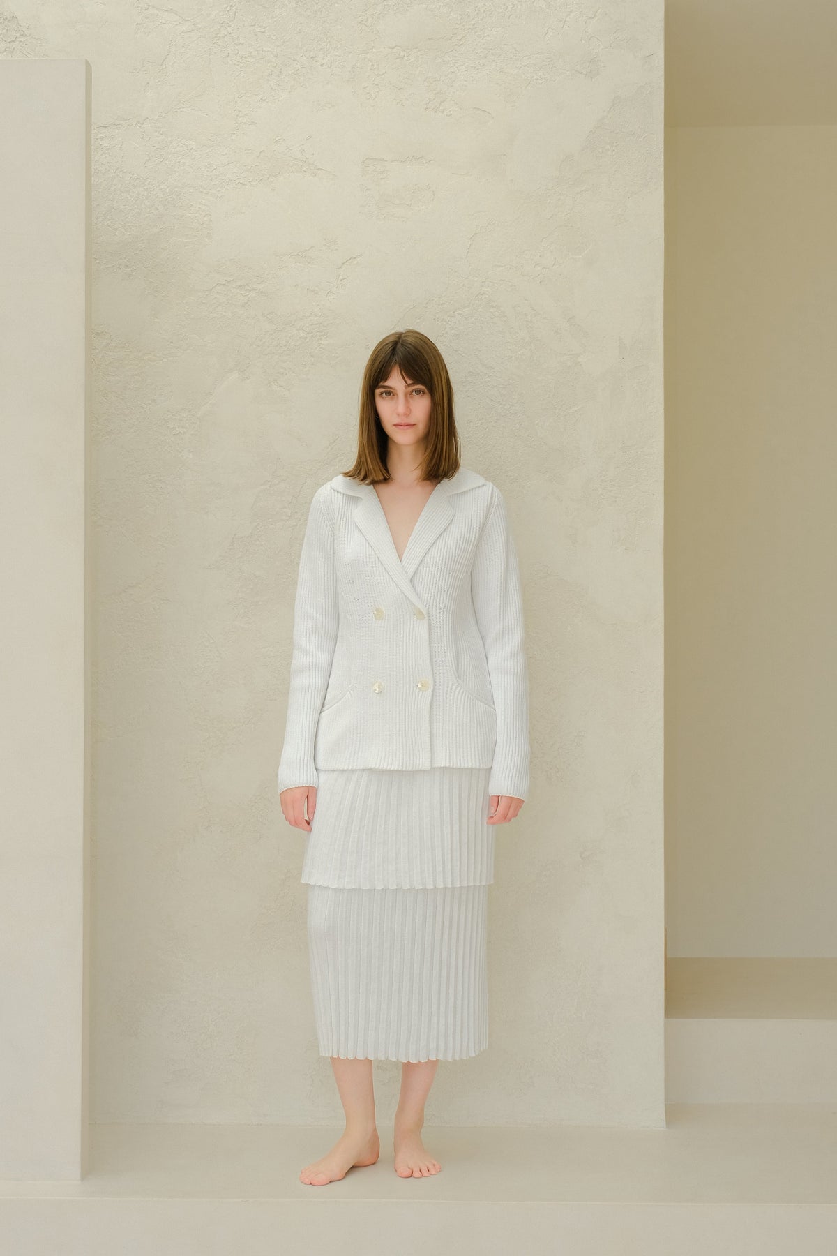 Modelo con a light blazer de punto cruzado and pale falda larga plisada against a neutral studio wall.