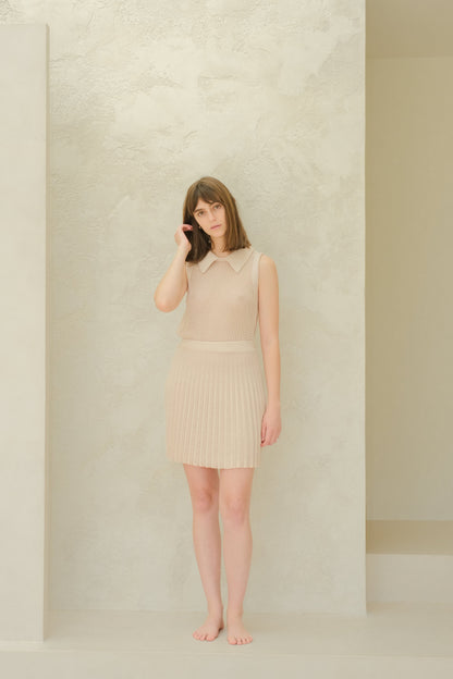 Modelo con a beige fine cotton plissé minifalda and top de punto sin mangas against a pale textured wall.