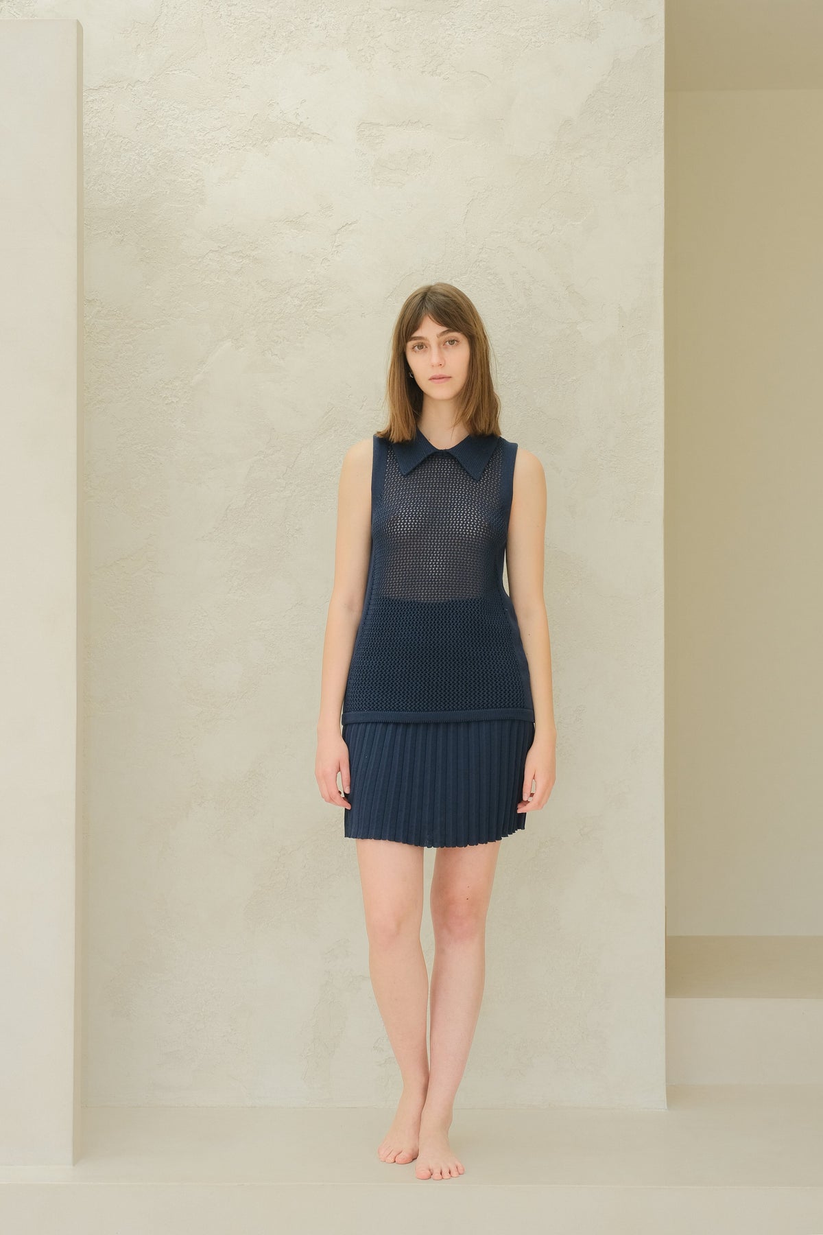 Vista frontal de cuerpo entero of a modelo con a azul marino sleeveless top de punto calado with a cuello puntiagudo and a pleated minifalda.