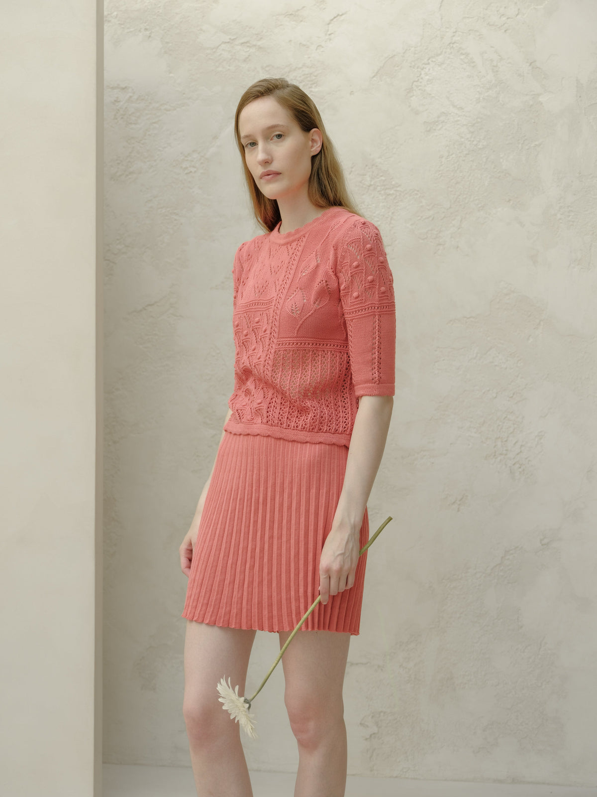Modelo con pink minifalda plisada de algodón fino with a top de punto a juego scuerods against a pale textured wall, sosteniendo una flor.