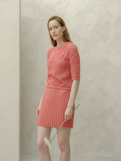 Modelo con pink minifalda plisada de algodón fino with a top de punto a juego scuerods against a pale textured wall, sosteniendo una flor.