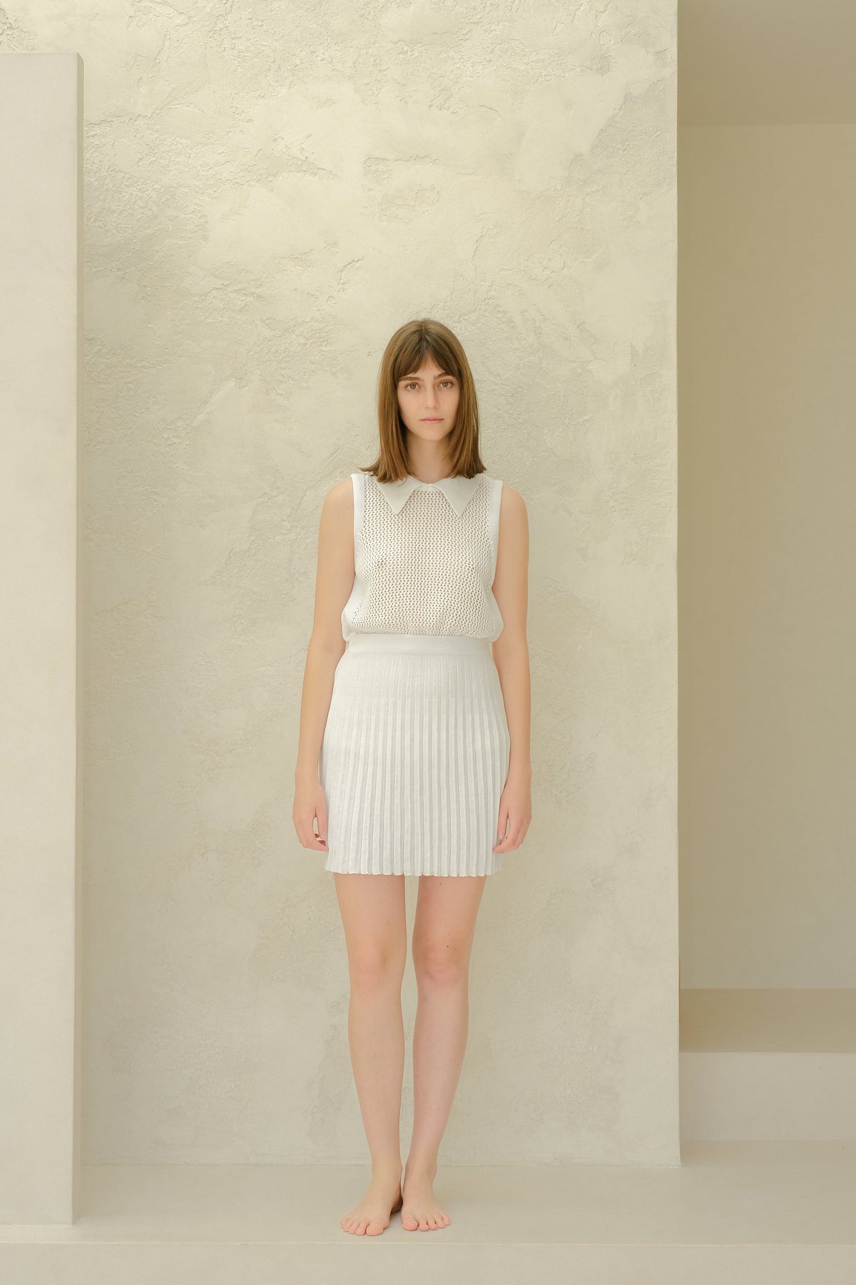 Vista de cuerpo entero of a modelo con a white mesh sleeveless top with a collared neckline and a white plissé minifalda, barefoot.