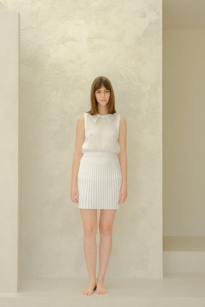 Vista de cuerpo entero of a modelo con a white mesh sleeveless top with a collared neckline and a white plissé minifalda, barefoot.