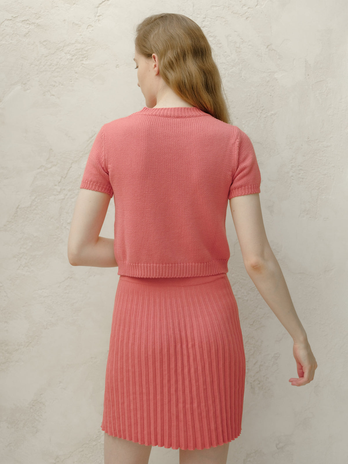Back view of a modelo con a coral pink knitted top and matching pleated minifalda