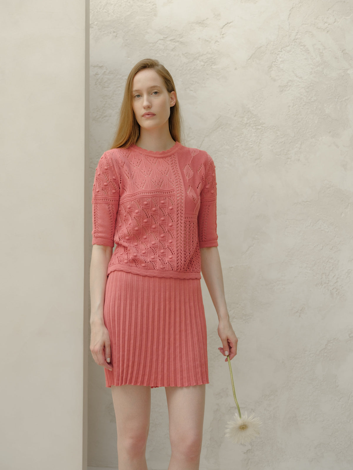 Vista frontal of a model in a pink minifalda plisada de algodón fino and top de punto a juego against a light textured wall.