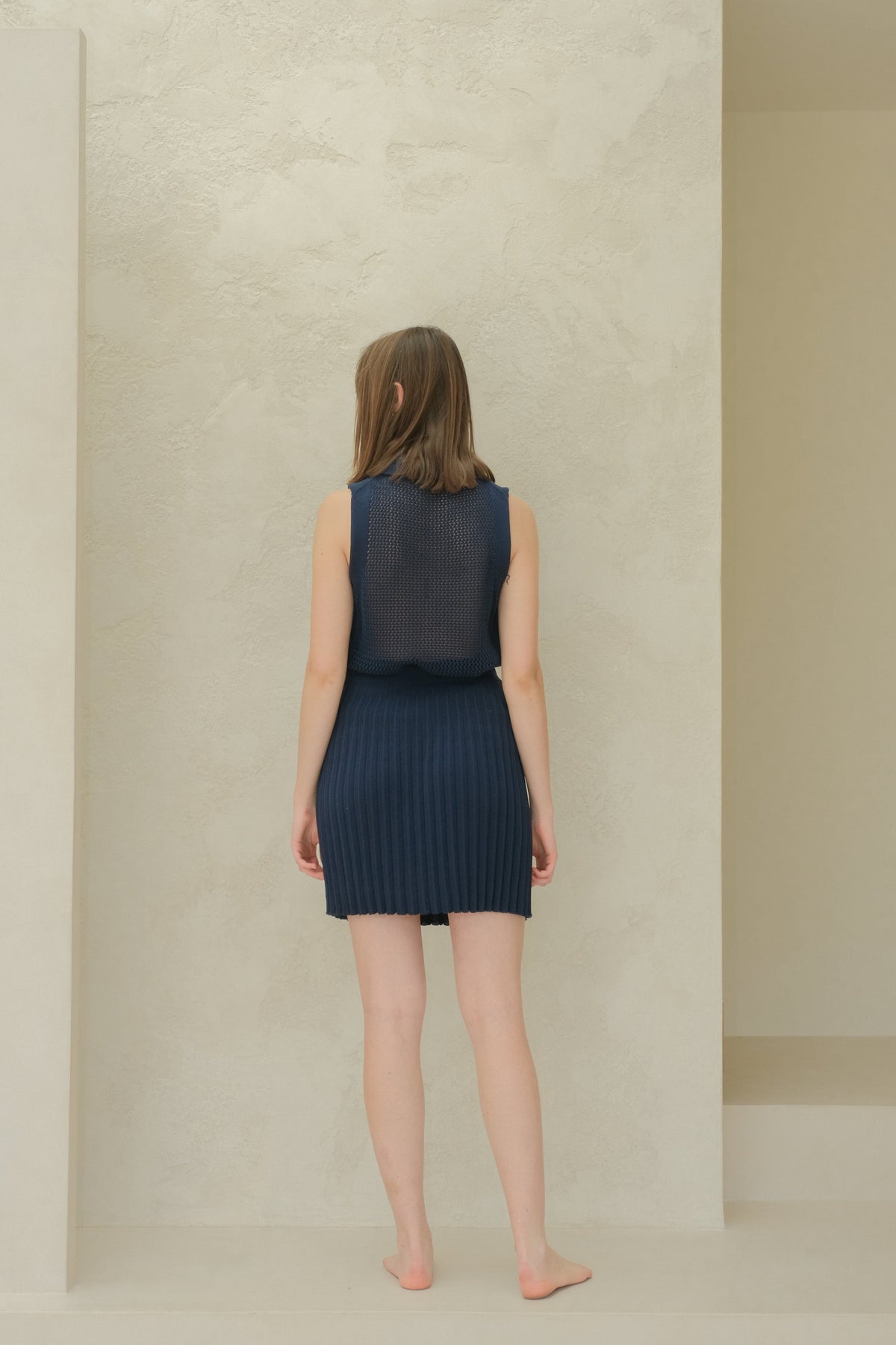 Back view of a model in a azul marino sleeveless top de punto calado with a cuello puntiagudo and pleated minifalda.