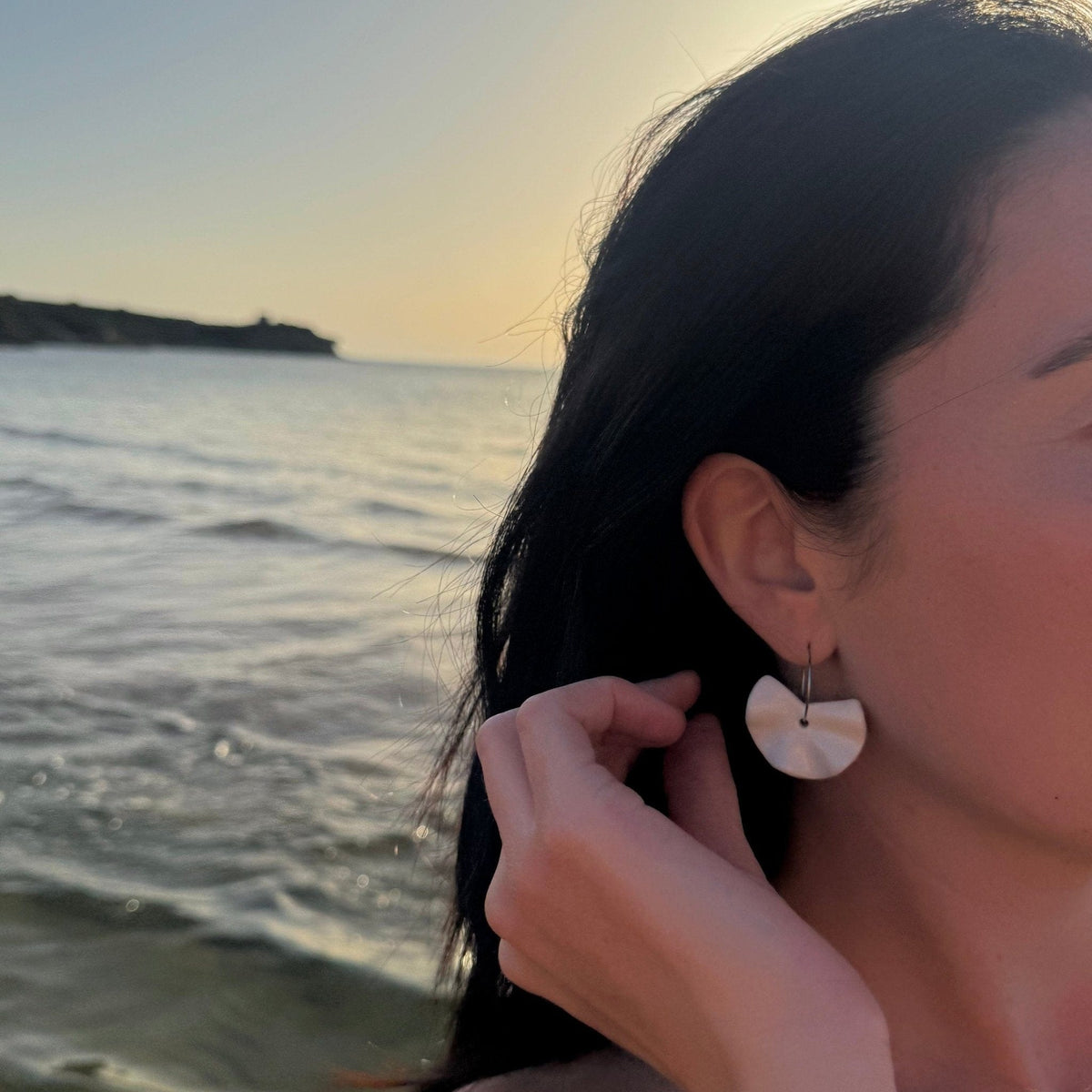 Primer plano of a mujer con Frolla white ceramic dangle earrings by the sea at sunset