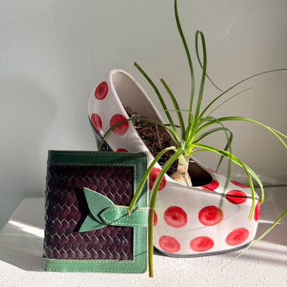 Lumi - Handwoven Leather Wallet | Green&Dark Coffee with woven dark coffee leather and green leather trim beside a white pot with red dots and plant.