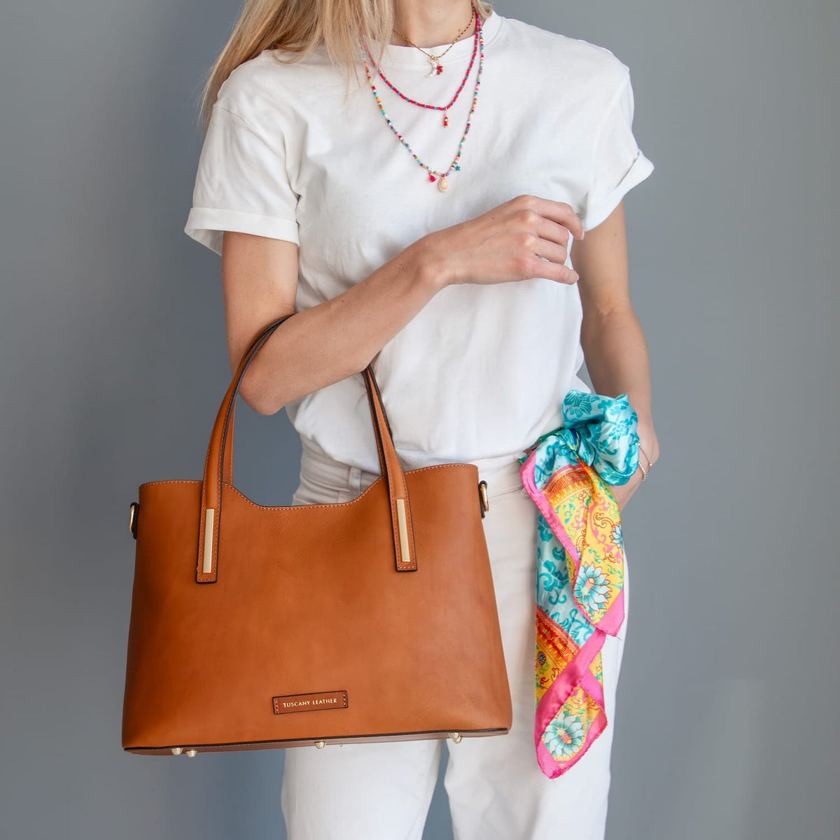 Person holding a brown leather tote handbag with a colorful scarf draped over their arm against a gray background