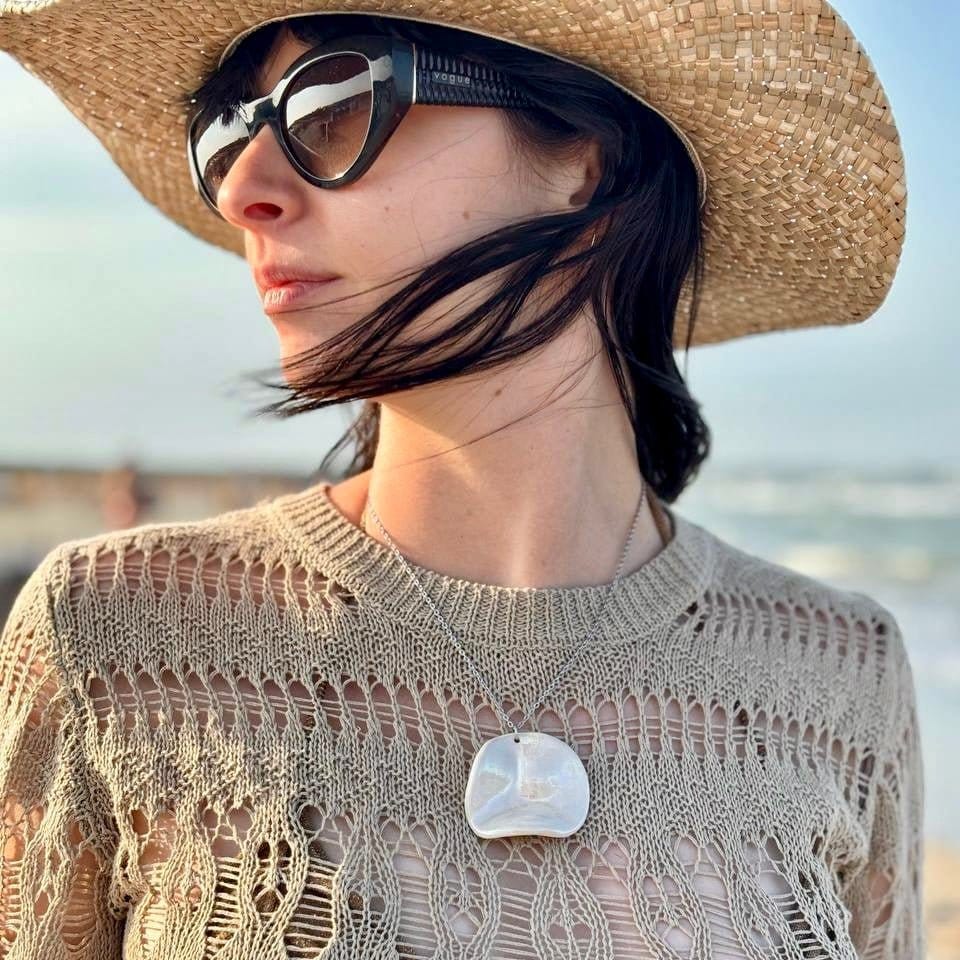 Woman wearing a large handcrafted pearl-white cermic necklace with a straw hat and sunglasses with a blurred beach background