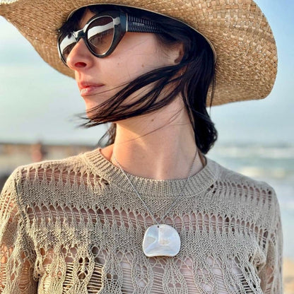 Woman wearing a large handcrafted pearl-white cermic necklace with a straw hat and sunglasses with a blurred beach background