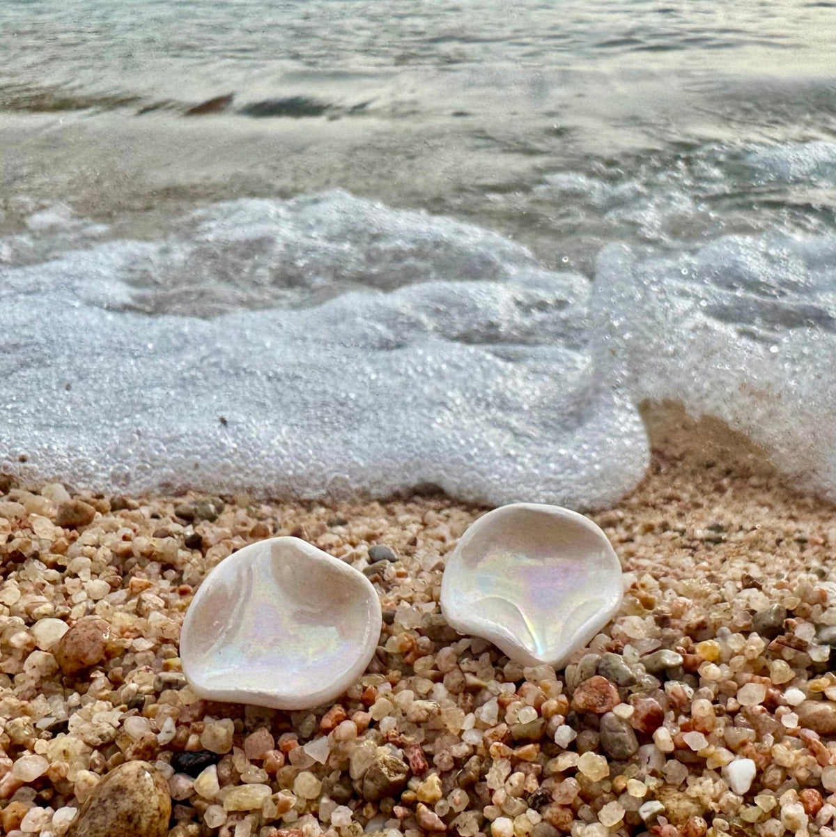 Deux boucles d’oreilles irisées en forme de coquillage sur une plage de galets avec des vagues en arrière-plan