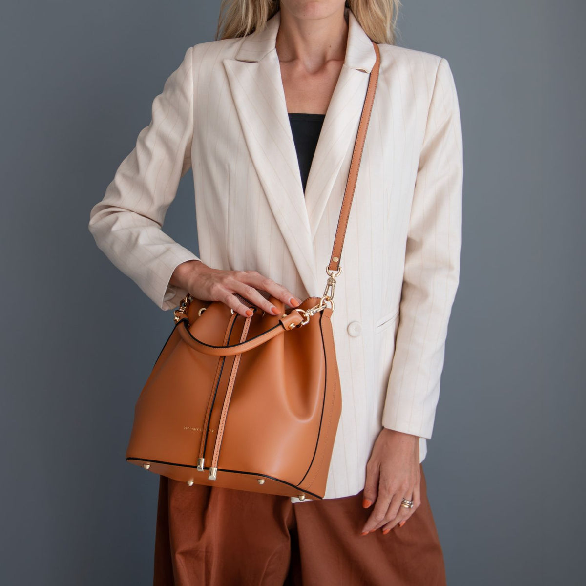 A donna con a crema blazer e marrone pants carries a cuoio leather bucket bag con a matching shoulder strap e accessori metallici color oro against a dark grigio studio background.