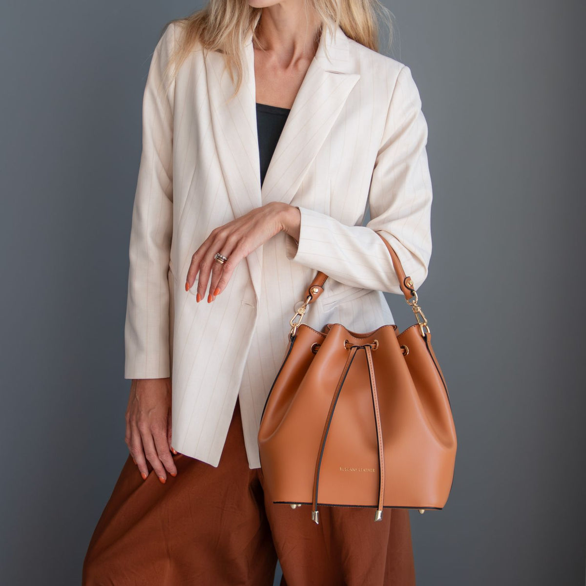 A modella con a crema blazer e marrone trousers carries a cuoio leather drawstring bucket bag con accessori metallici color oro against a dark grigio studio background.