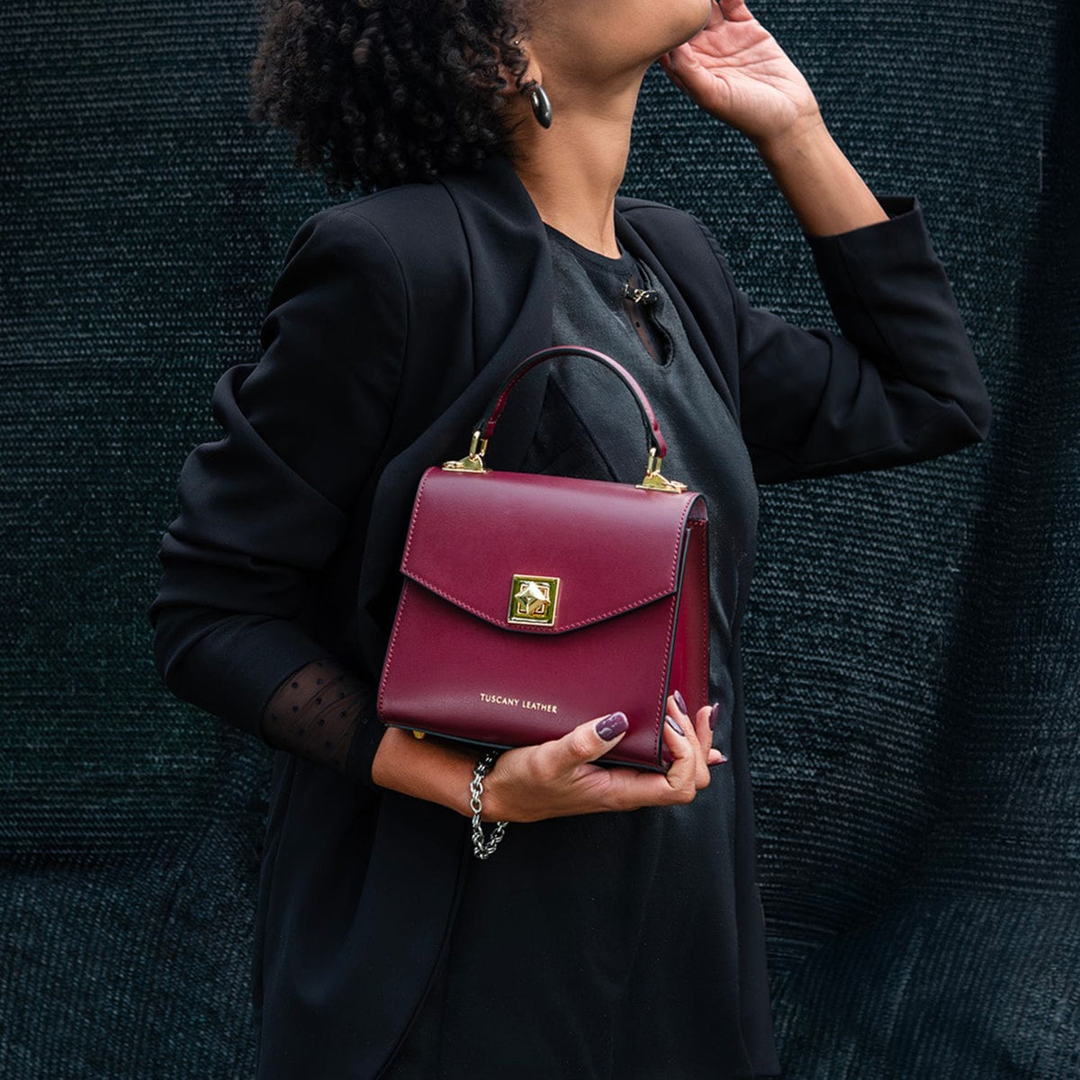 Burgundy leather TL Bag mini bolso with a cierre cuadrado dorado and asa superior, sostenido por una persona in a black blazer.